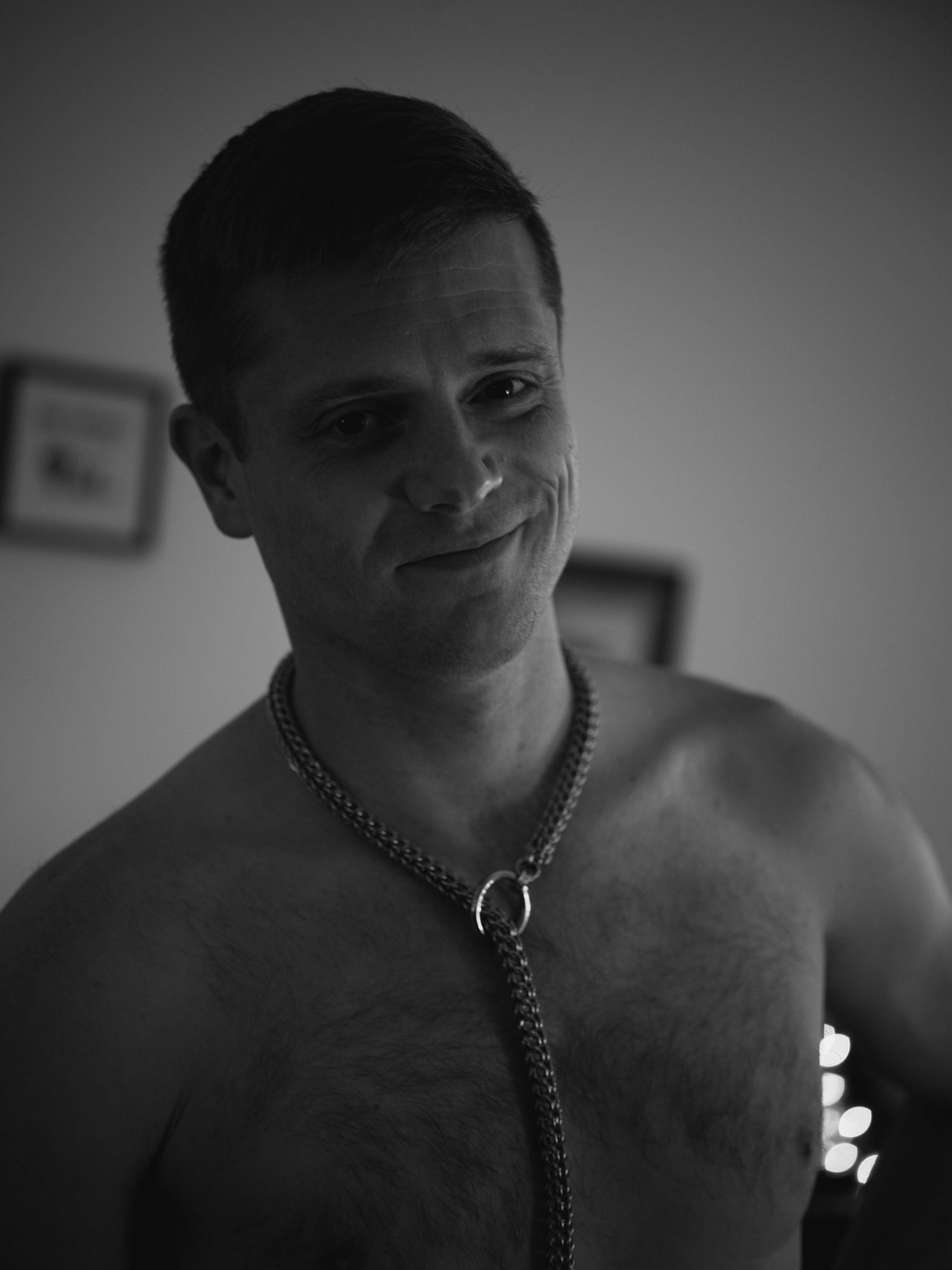 A black and white portrait photograph of the artist, Nolan Frame, wearing The Good Boy Stainless Steel Zeke necklace on his shirtless and hairy male torso. Nolan is smirking directly at the camera, with shadow lightly obscuring one side of his face.